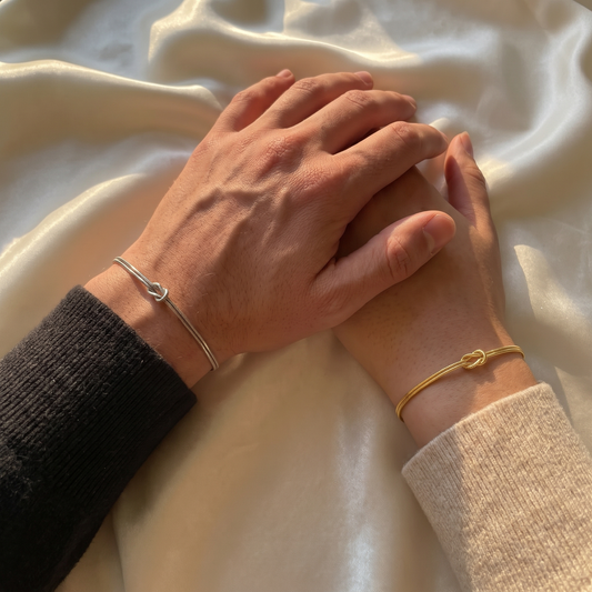 Two hands resting together on silk, one wearing the silver knot bracelet and one the gold