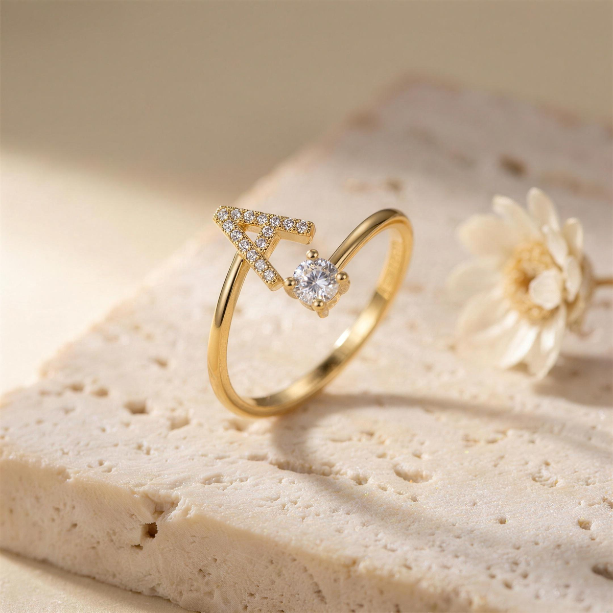 Gold open ring with a paved letter A and a round zirconia on a travertine slab beside a dried flower