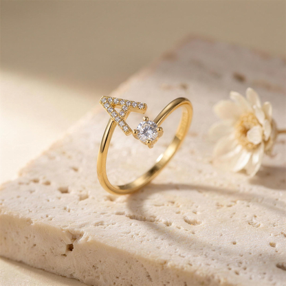 Gold open ring with a paved letter A and a round zirconia on a travertine slab beside a dried flower