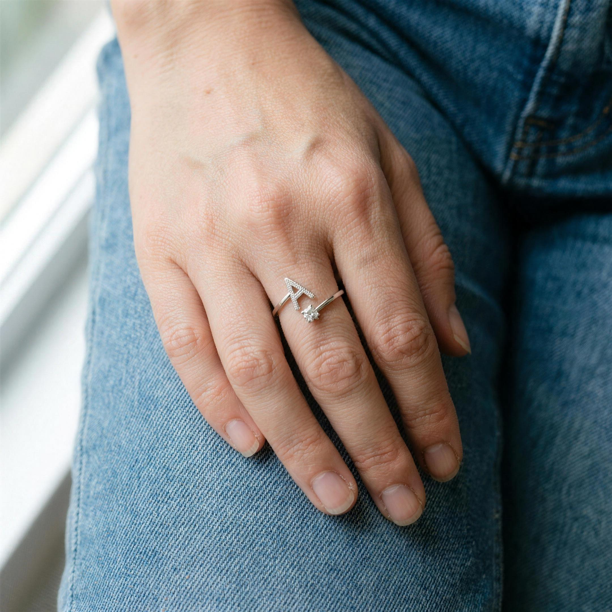 Silver initial ring with a paved letter A worn on the ring finger, hand resting against blue jeans