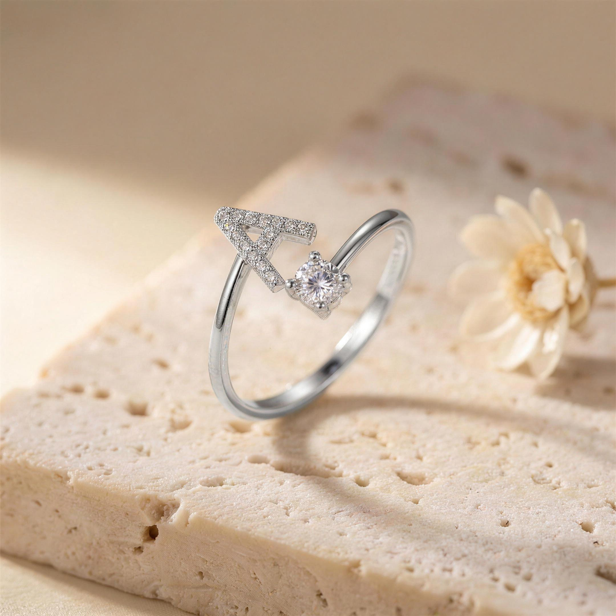 Silver open ring with a paved letter A and a round zirconia on a travertine slab beside a dried flower