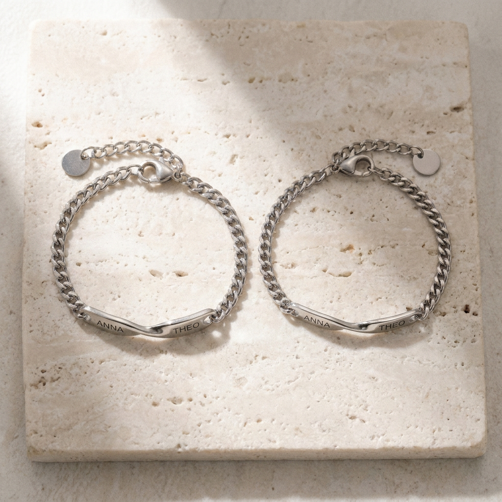 The two matching name bracelets, closed, side by side on pale travertine stone, ANNA and THEO readable