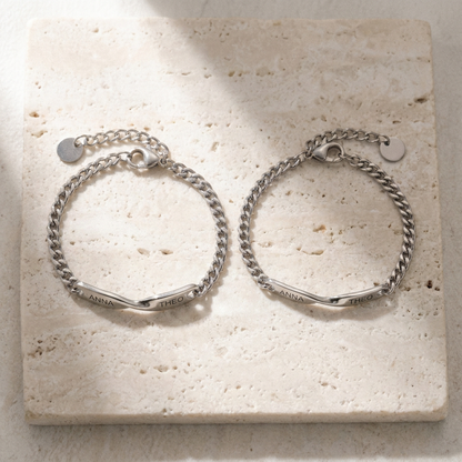The two matching name bracelets, closed, side by side on pale travertine stone, ANNA and THEO readable