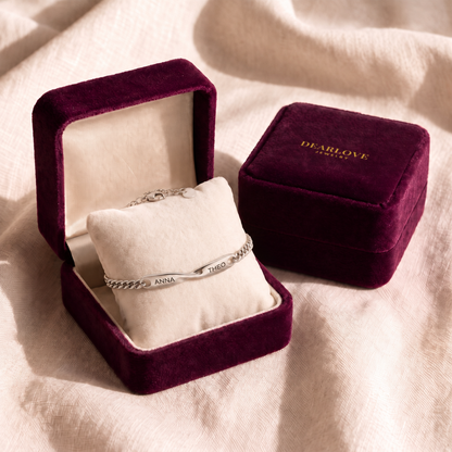 A plum velvet gift box open on cream linen with the matching name bracelet on its cushion, the closed box beside it stamped DEARLOVE JEWELRY in gold