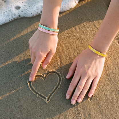 Hands drawing a heart in the sand, wearing sea green, soft pink and yellow cuffs
