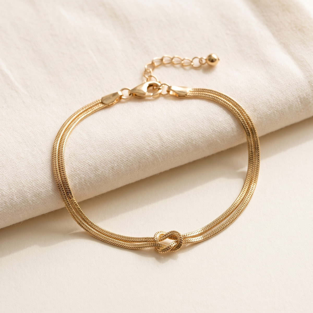 The gold Forever Knot bracelet open on cream cotton, clasp and extender chain visible