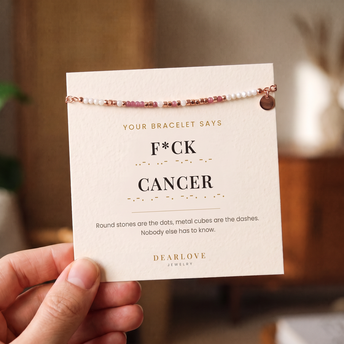 Hand holding the bracelet on its card, which reads F*ck Cancer in Morse code