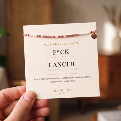 Hand holding the bracelet on its card, which reads F*ck Cancer in Morse code