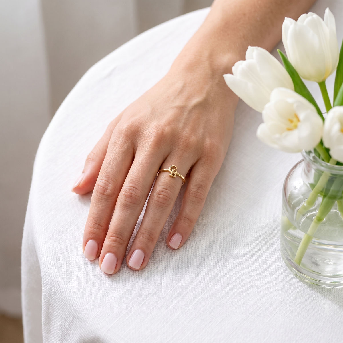 The gold interlocking initial ring worn on a hand resting beside white tulips