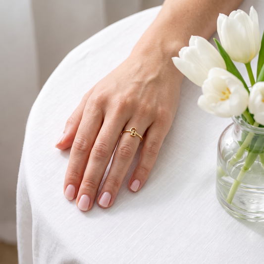 The gold interlocking initial ring worn on a hand resting beside white tulips