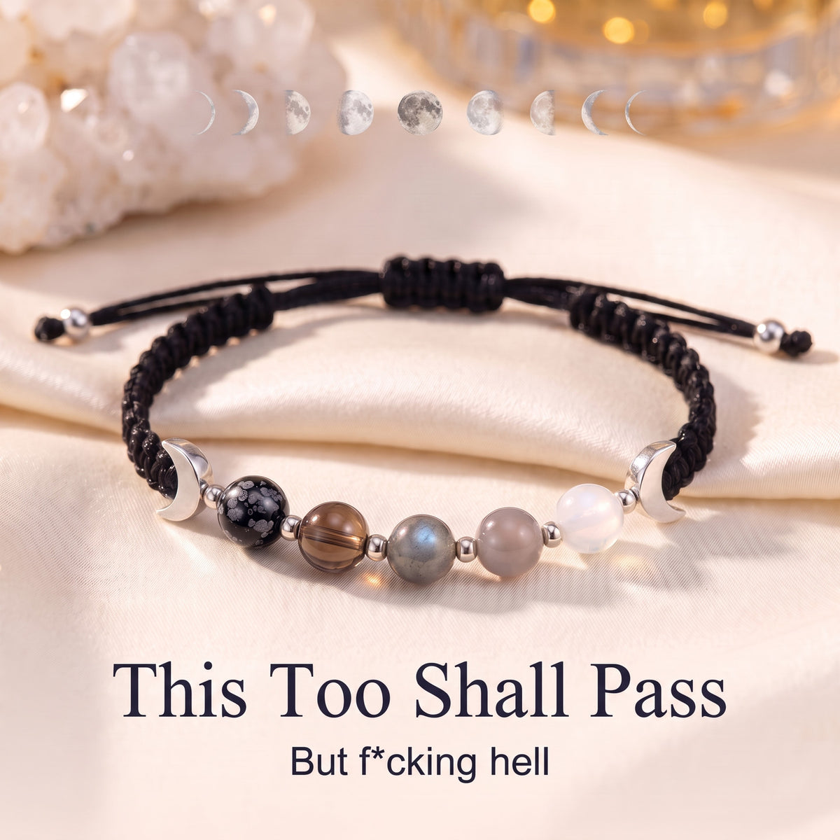 The bracelet on ivory satin under a line of moon phases, with the words This Too Shall Pass and But f*cking hell