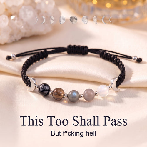 The bracelet on ivory satin under a line of moon phases, with the words This Too Shall Pass and But f*cking hell
